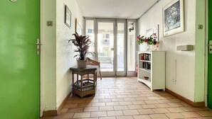 A hallway with a green door, a wicker basket with plants, a wicker chair, a white cabinet with books, and a tiled floor. - Cozy F2 Apartment, A/C, Terrace, Near Thermes & Park, Pet-Friendly, Lamalou-les-Bains (Lamalou-les-Bains)