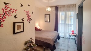 Un dormitorio con cama, mesita de noche con lámpara, ventana con cortinas, pared decorada con motivos florales y aves, y una luz en el techo.