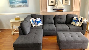 A modern living room with a sectional sofa and ottoman, a small side table, and framed nautical-themed artwork on the wall. - Best Beachfront - Expansive OCEAN VIEWS Perfect location! 2 decks & large patio (Ocean City)