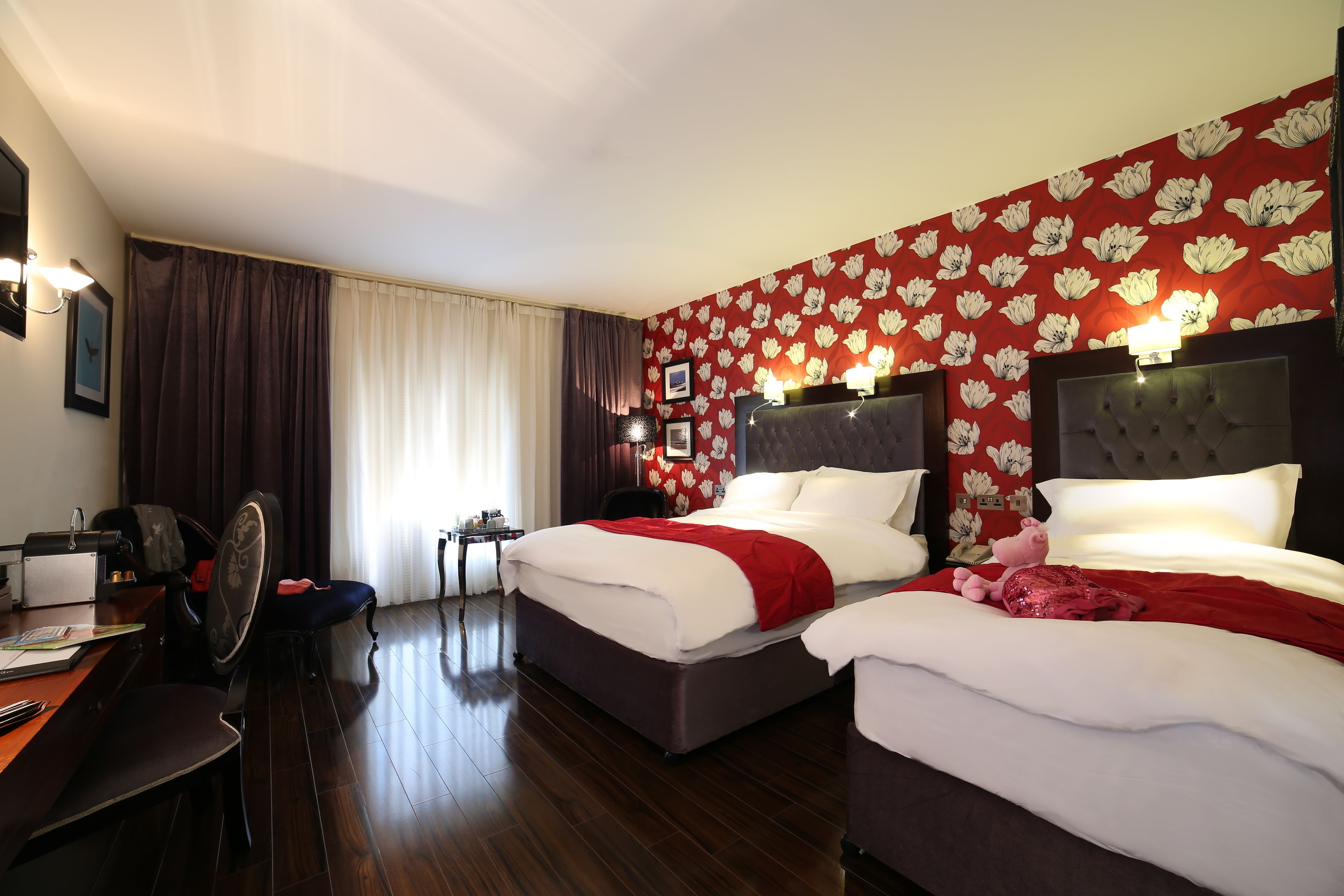 Una camera d'albergo con due letti, una scrivania, una sedia e un'ampia finestra con tende.