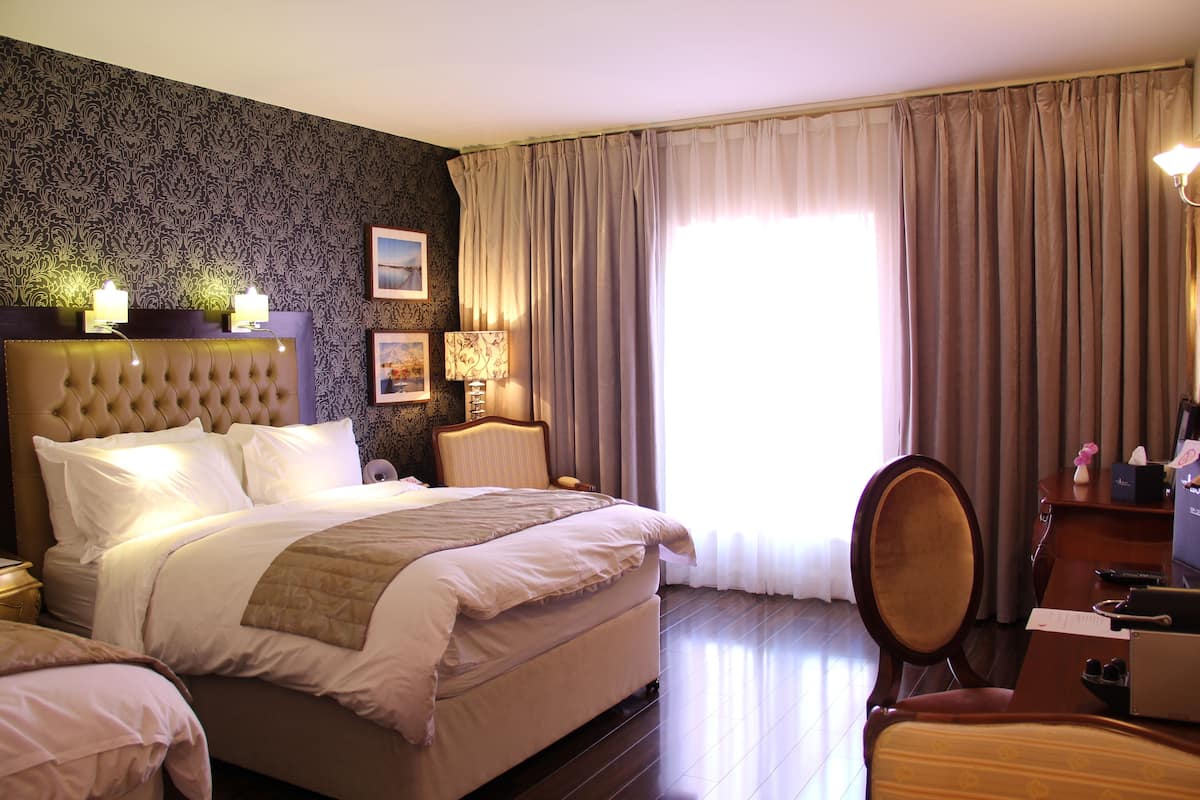 Quarto de hotel com uma cama grande, uma mesa com cadeira, uma luminária e uma janela com cortinas.