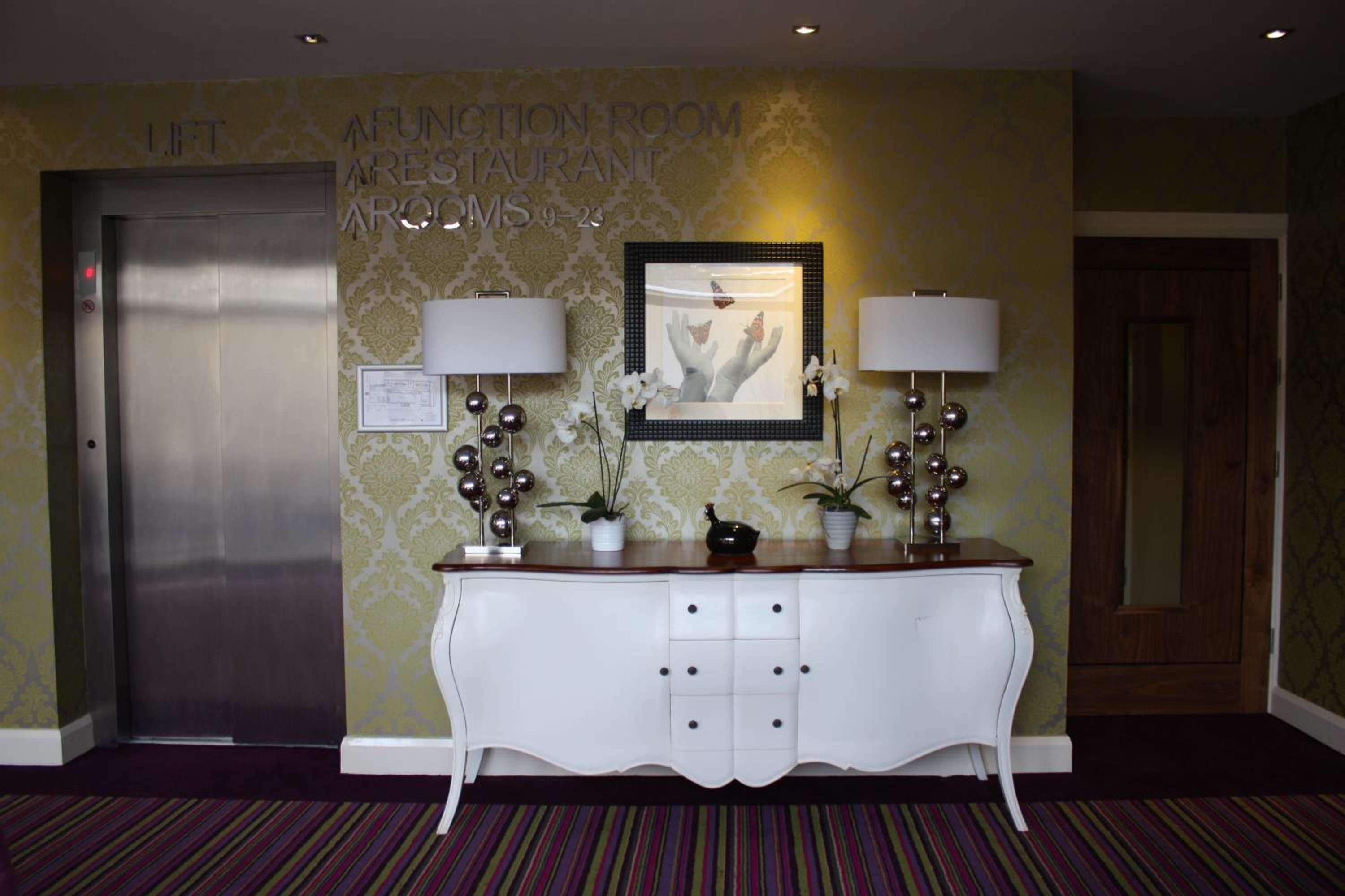 An elevator, a reception desk with two lamps, a framed picture, and a wall with a sign indicating the location.