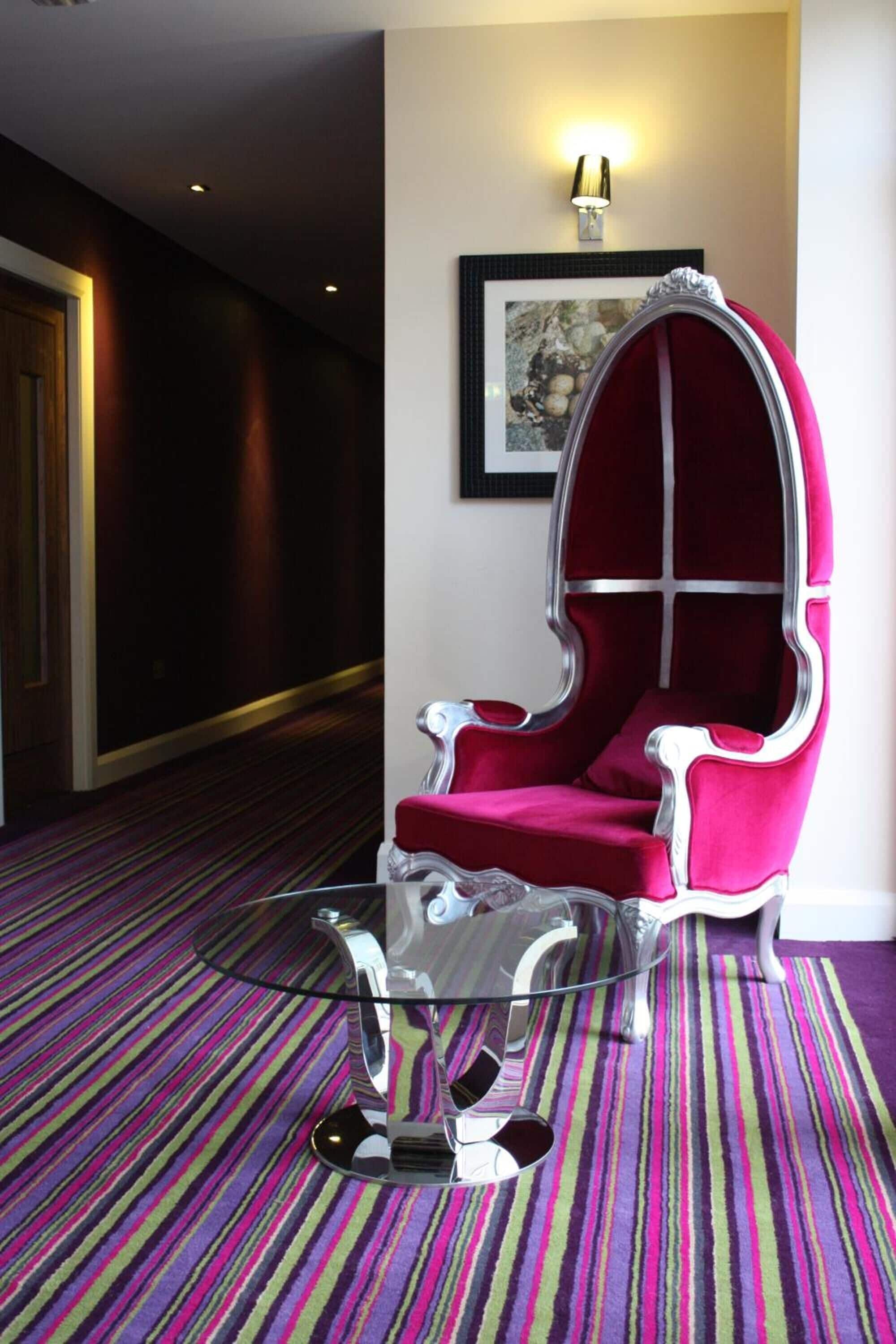 A modern hotel room with a striped carpet, a clear glass coffee table, and a red upholstered chair.