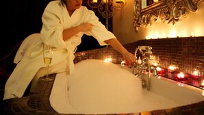 A person in a white bathrobe leaning over a bathtub with a glass of wine, candles, and a mirror in the background. - Talbot Hotel (Belmullet)