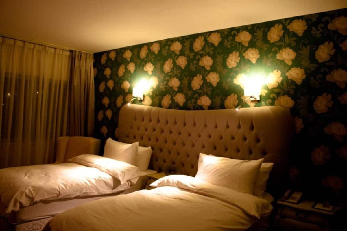 Quarto de hotel com duas camas, cabeceira estofada, papel de parede floral e duas luminárias fixadas na parede.