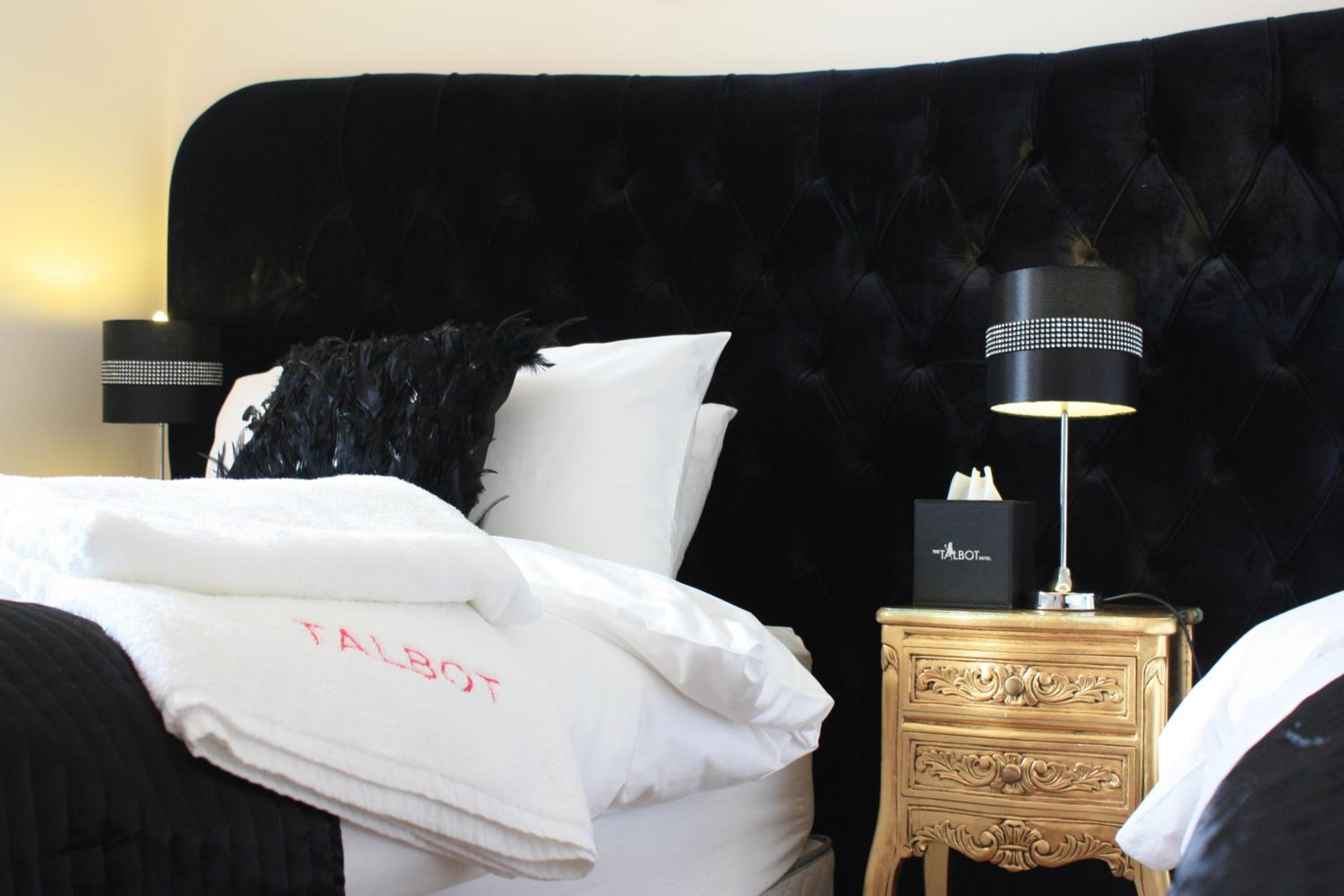 Een bed met witte lakens, een zwart veren kussen, een Talbot handdoek, een zwarte lamp en een gouden nachtkastje met een doos.