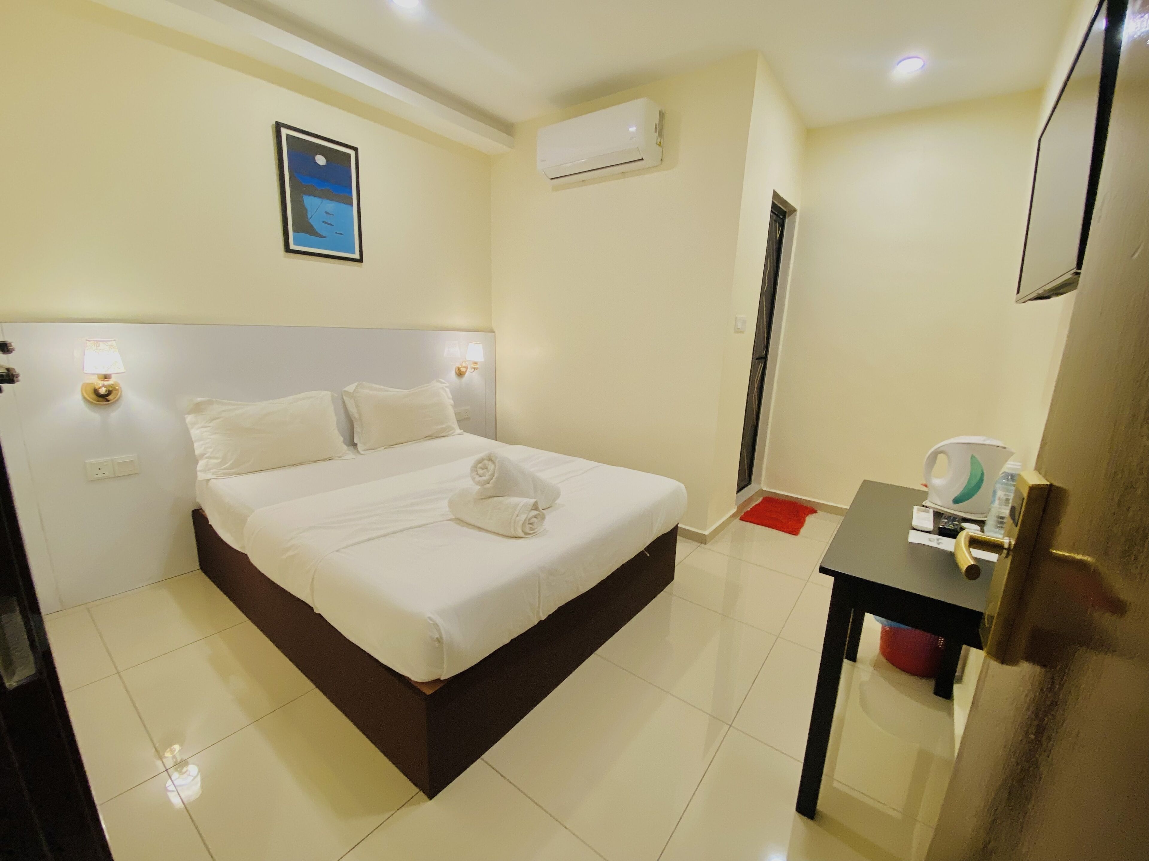 A hotel room with a bed, a bedside table, an air conditioning unit, a framed picture on the wall, and a red rug on the floor.