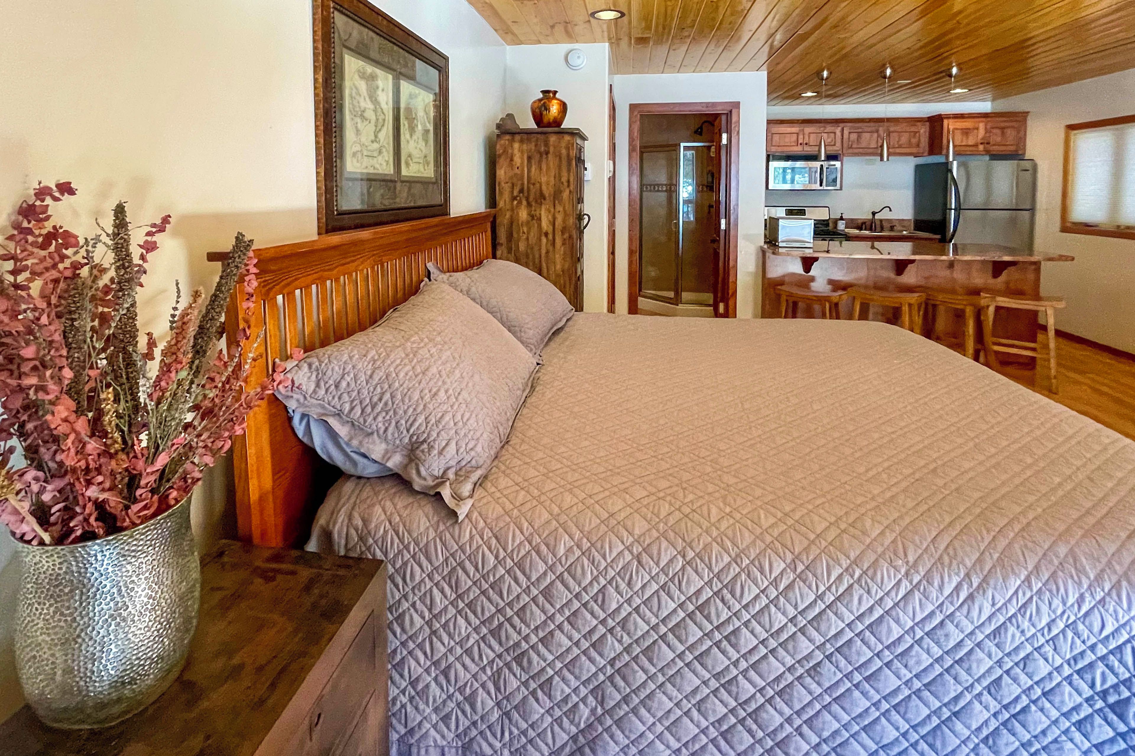 Une chambre avec un grand lit, une tête de lit en bois, une table de chevet ornée d’un vase de fleurs, et une vue sur un coin cuisine équipé d’un four à micro-ondes et d’un évier.