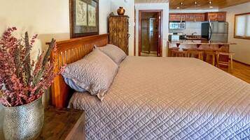 A bedroom with a large bed, a wooden headboard, a bedside table with a vase of flowers, and a view into a kitchen area with a microwave and a sink.