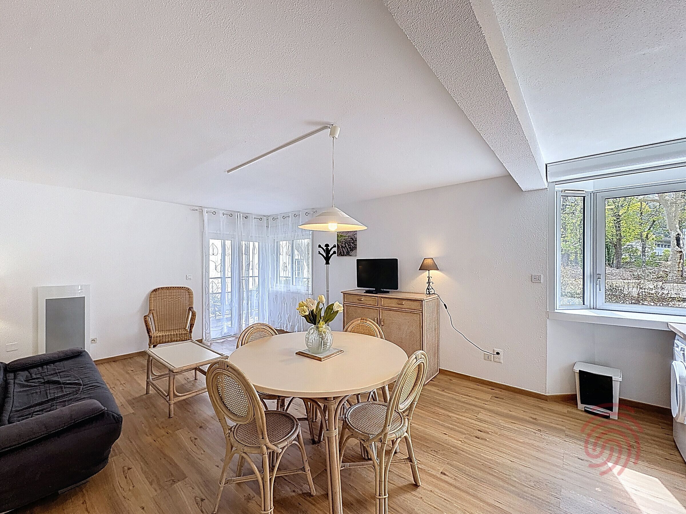 A modern living room with a round dining table, wicker chairs, a black sofa, a TV, and a window with a view of trees.