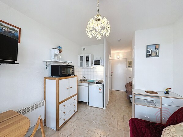 A compact kitchen with a microwave, toaster, and refrigerator. A wooden cabinet with drawers is present, and a small table with a chair is visible.