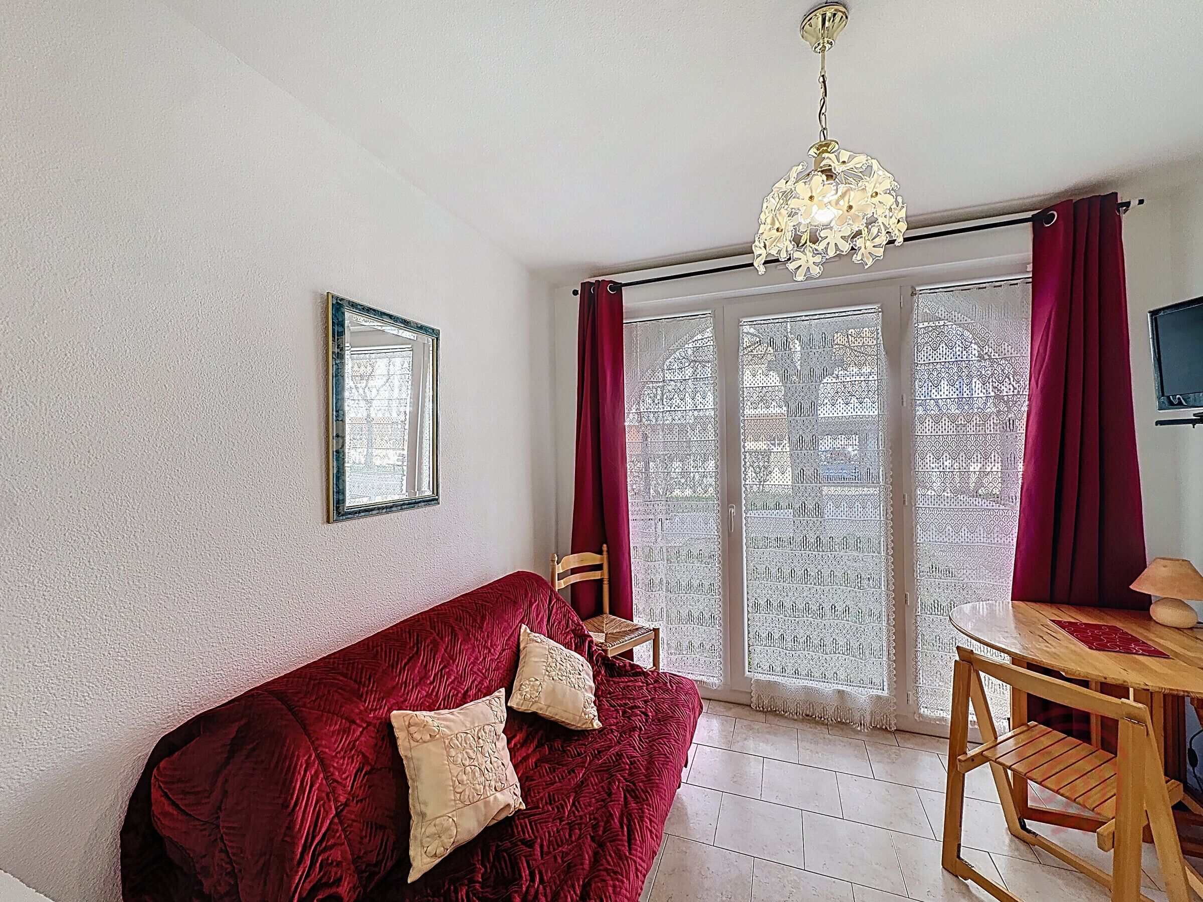 A room with a red sofa, a wooden chair, a table with a lamp, and a large window with patterned curtains.