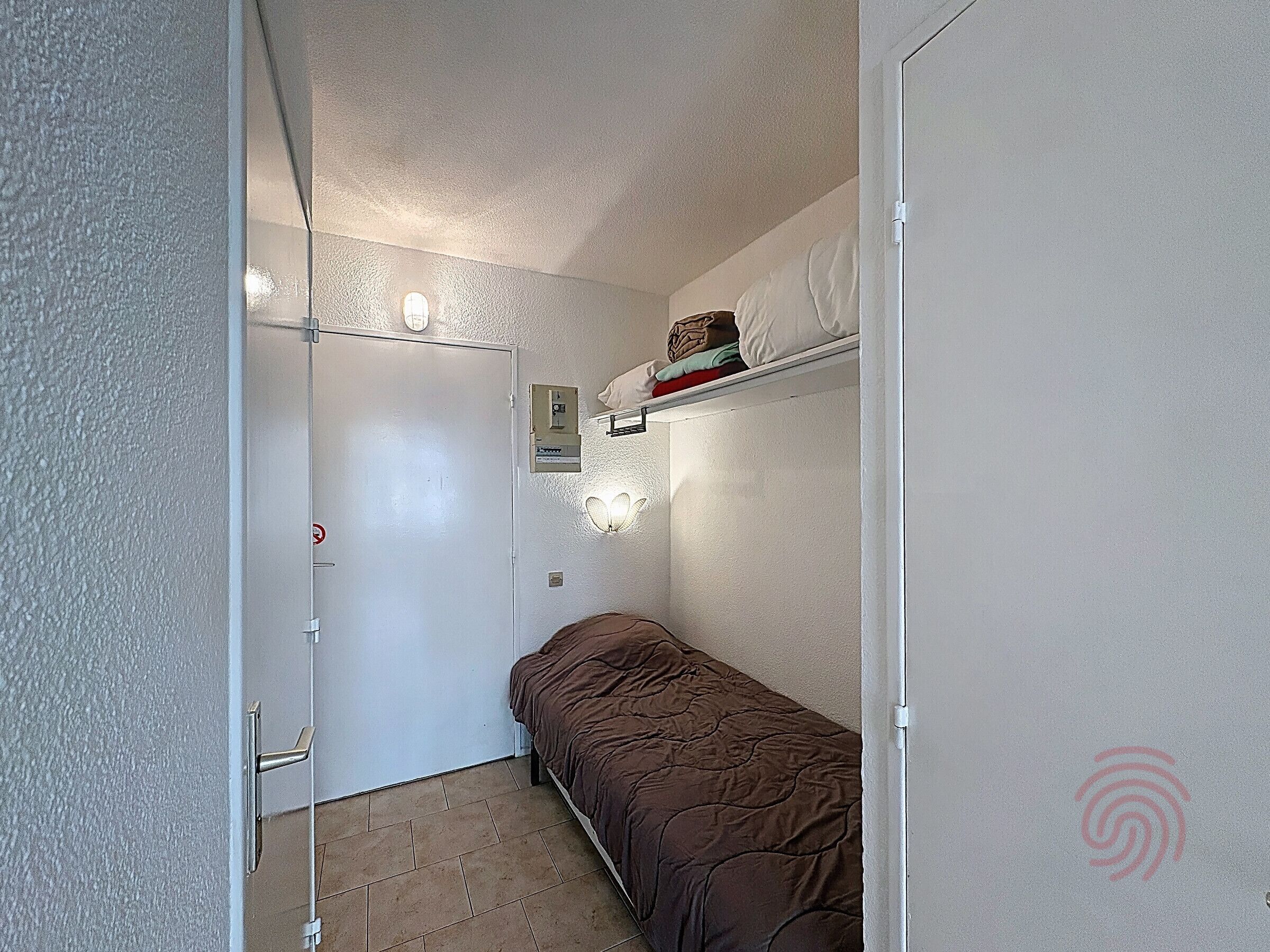 A small, narrow room with a single bed, a door, a shelf with folded clothes, and a wall-mounted light fixture.