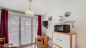 Ein Zimmer mit großem Fenster, einem Holzsessel, einem kleinen Tisch, einem Schrank, einer Mikrowelle, einem Fernseher und einer Deckenleuchte.