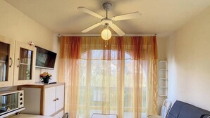Ein Zimmer mit Deckenventilator, Fernseher auf einem Schrank, einem Esstisch und einem Fenster mit durchsichtigen Vorhängen.