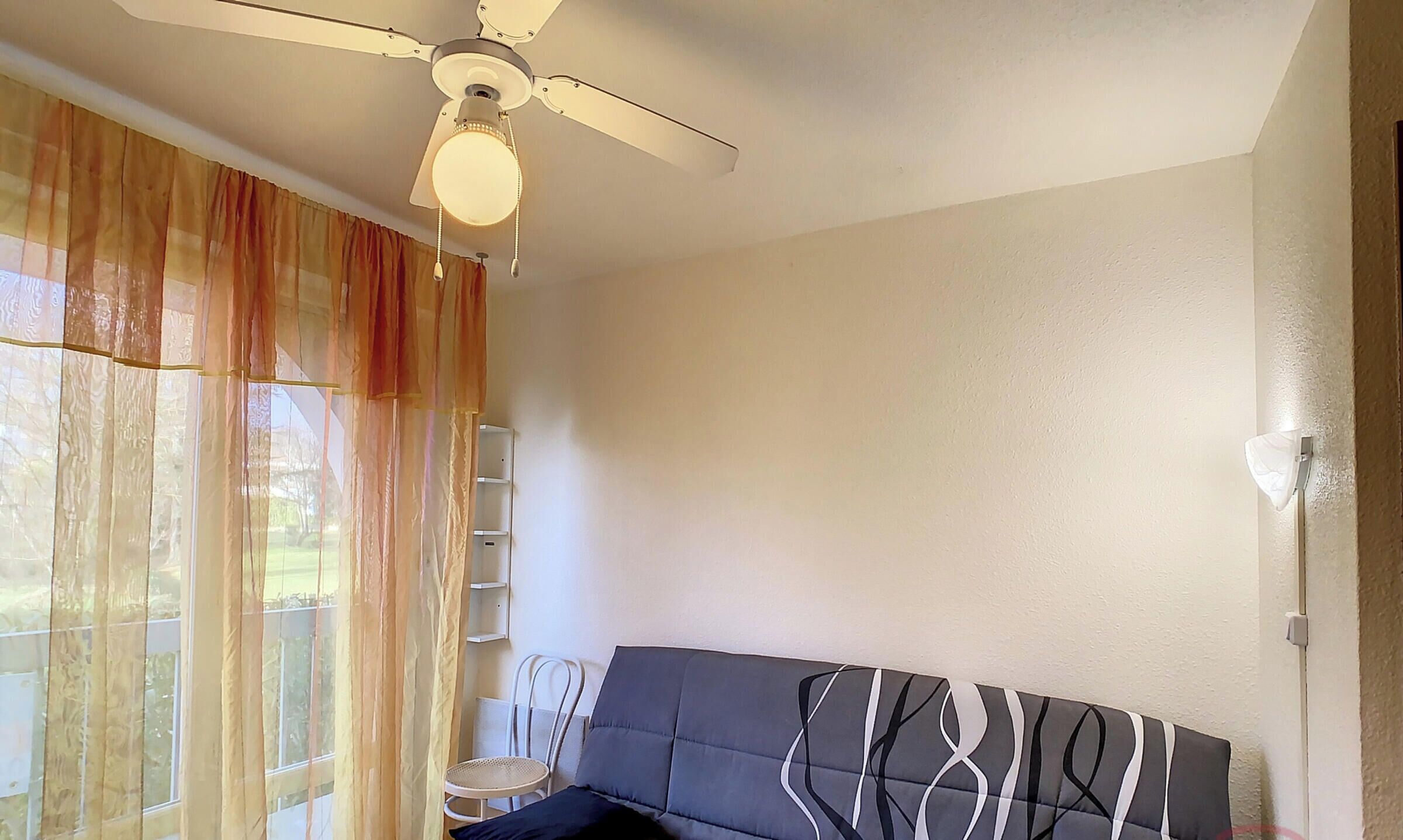 Una habitación con ventilador de techo, un sofá, una silla y una ventana con cortinas translúcidas.
