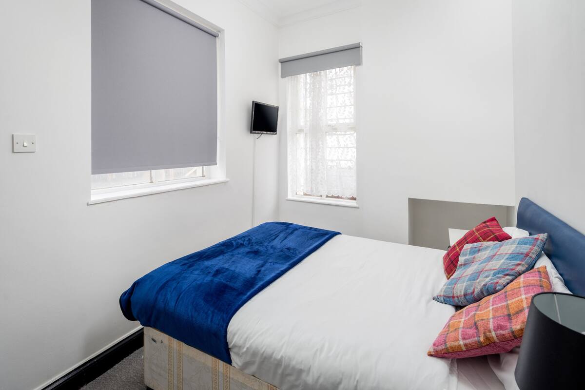 A bedroom with a bed, a television, a window with a blind, and a nightstand.