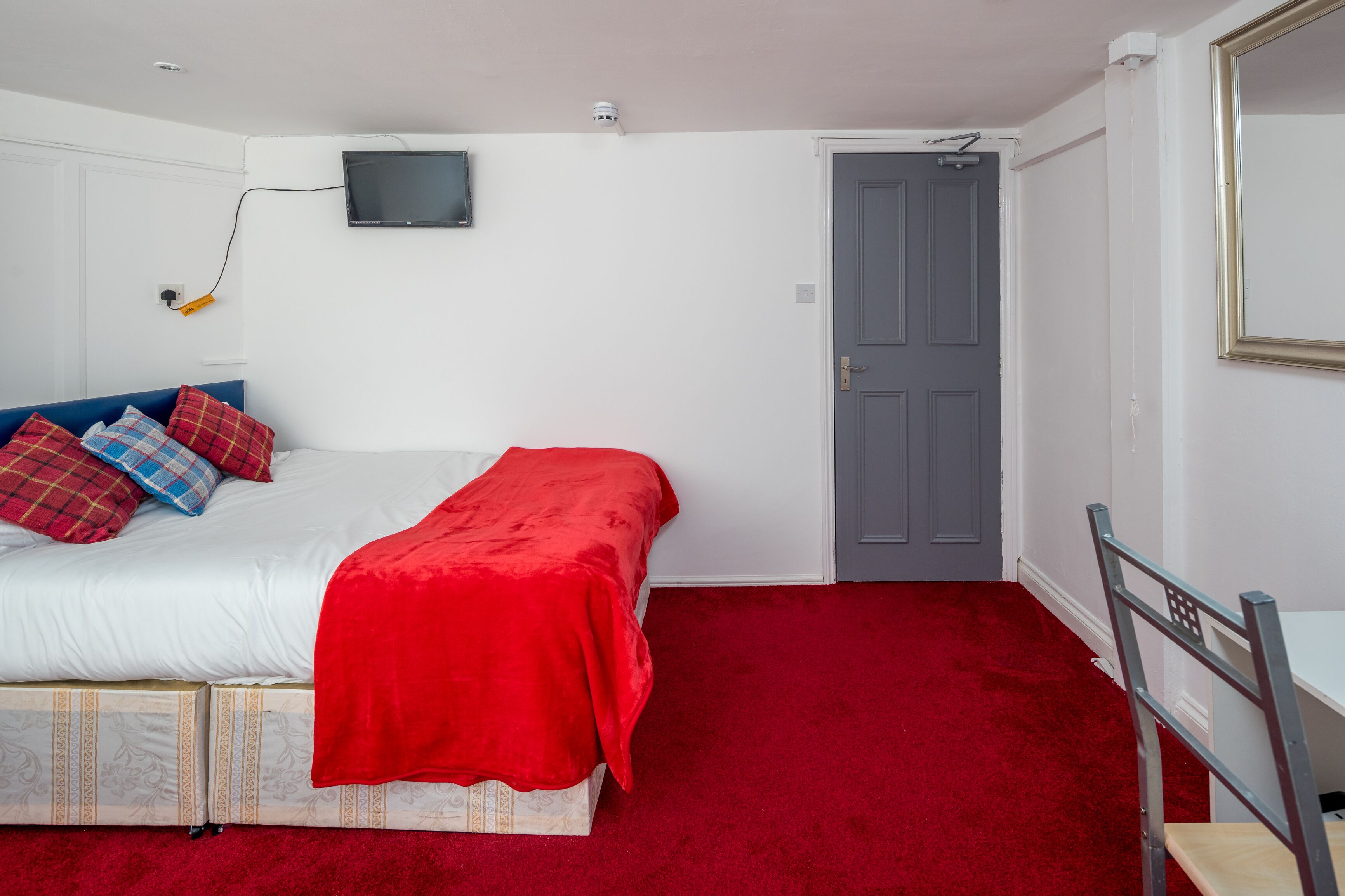 A bedroom with a bed, a red carpet, a grey door, and a television mounted on the wall.