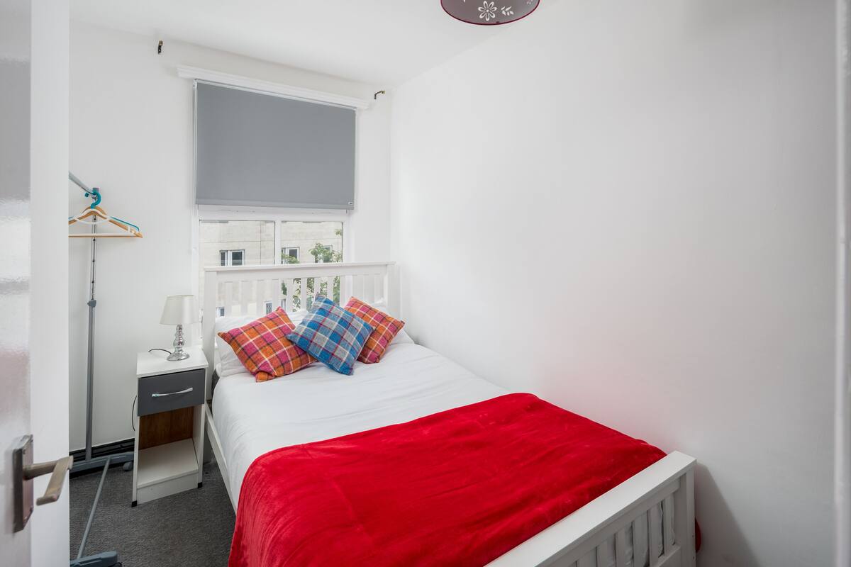 A bedroom with a white bed, a red blanket, a nightstand with a lamp, a window with blinds, and a white wardrobe.