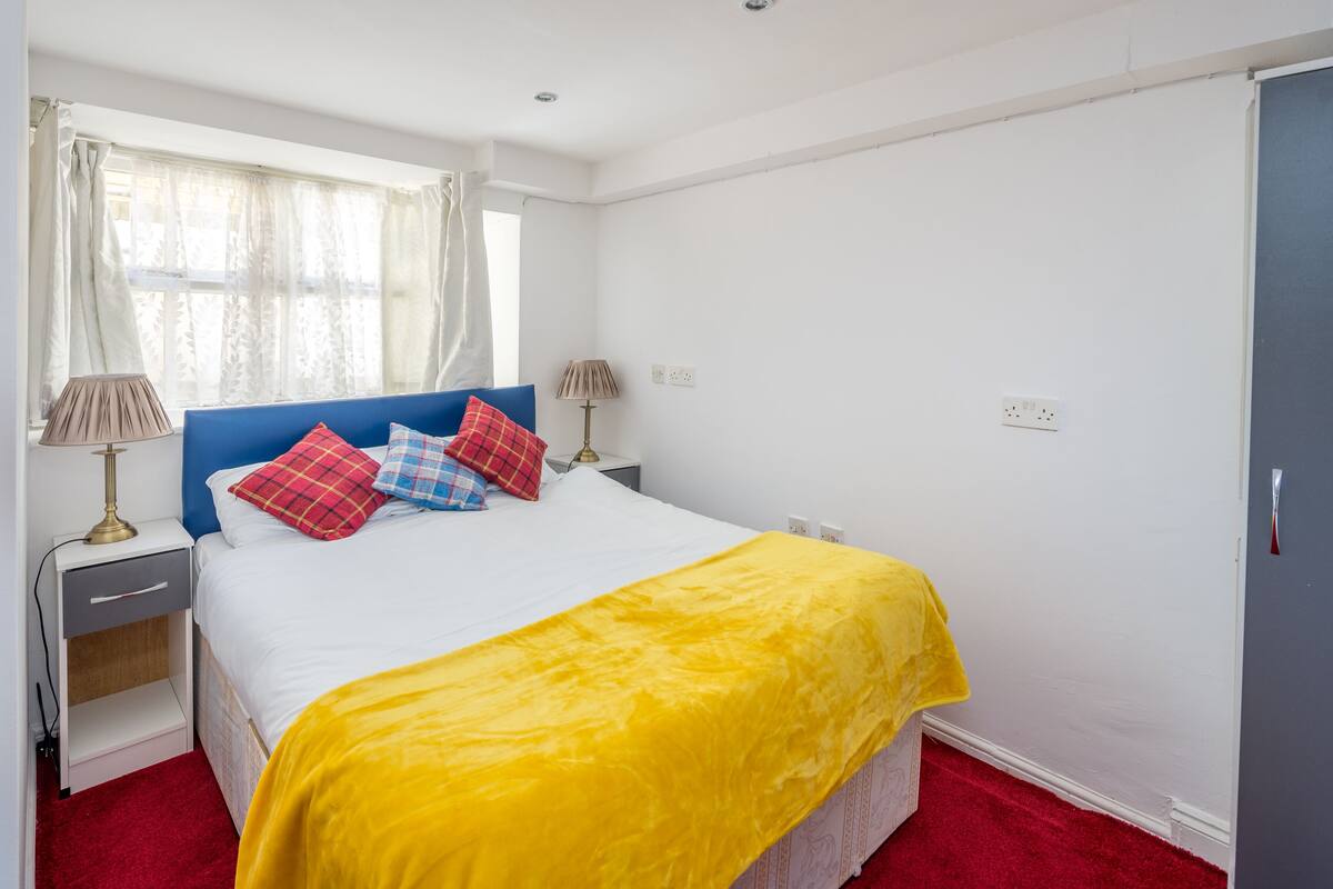 A bedroom with a bed, two bedside tables, a nightstand, a window with curtains, and a wardrobe.
