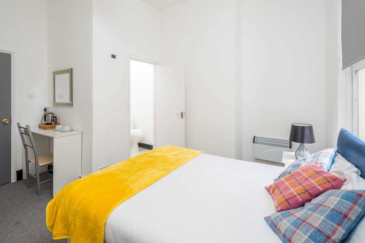 A hotel room with a bed, a desk, a chair, a mirror, a window, and a radiator.