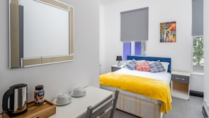 A hotel room with a bed, bedside table, a desk with a kettle, a coffee maker, and a large mirror.