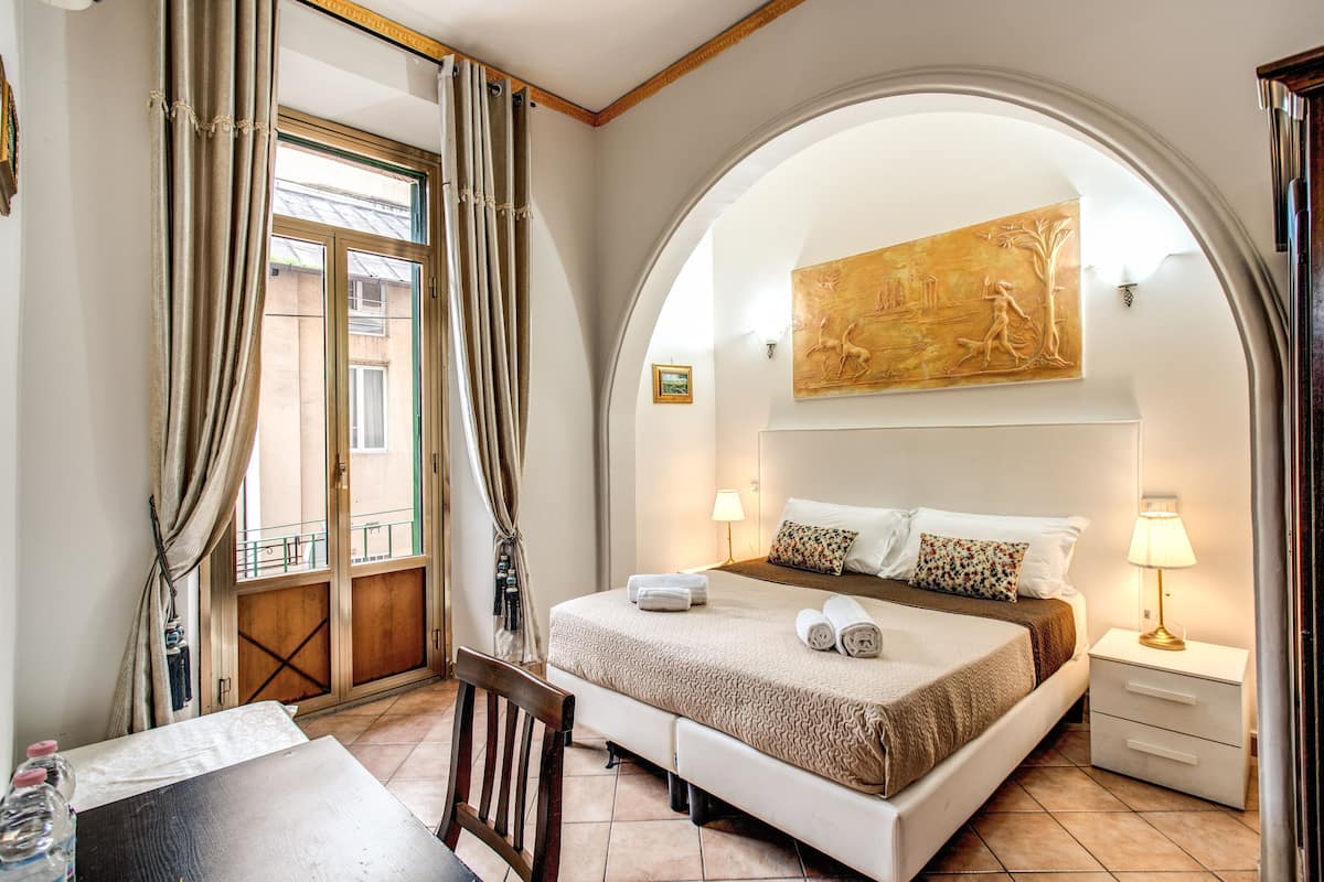 Una habitación de hotel con cama, escritorio, silla, mesita de noche y un cuadro en la pared.
