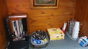 Una cafetera, un cesto de cápsulas de café, una caja de té Lipton y una pila de tazas blancas sobre una pared de madera.