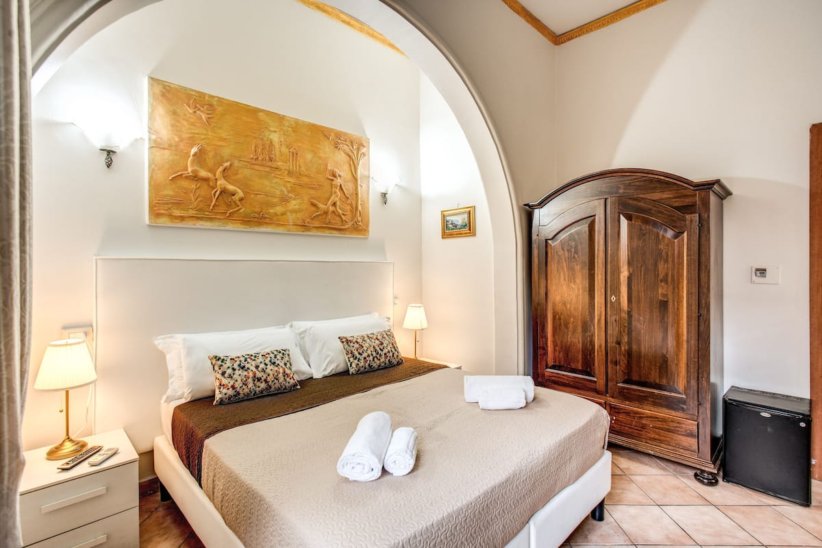 Una habitación de hotel con una cama, un armario de madera, un cuadro en la pared y una mesita de noche con una lámpara.