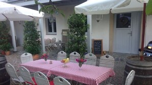 An outdoor dining area with a table set for six, surrounded by white metal chairs with red cushions. There are two umbrellas providing shade, and a chalkboard menu is visible on the wall. - Adele - Gästezimmer am Elberadweg (Dresden)