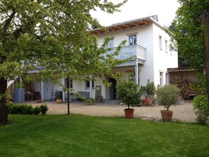 A two-story house with a balcony, surrounded by a garden with potted plants and a lawn.