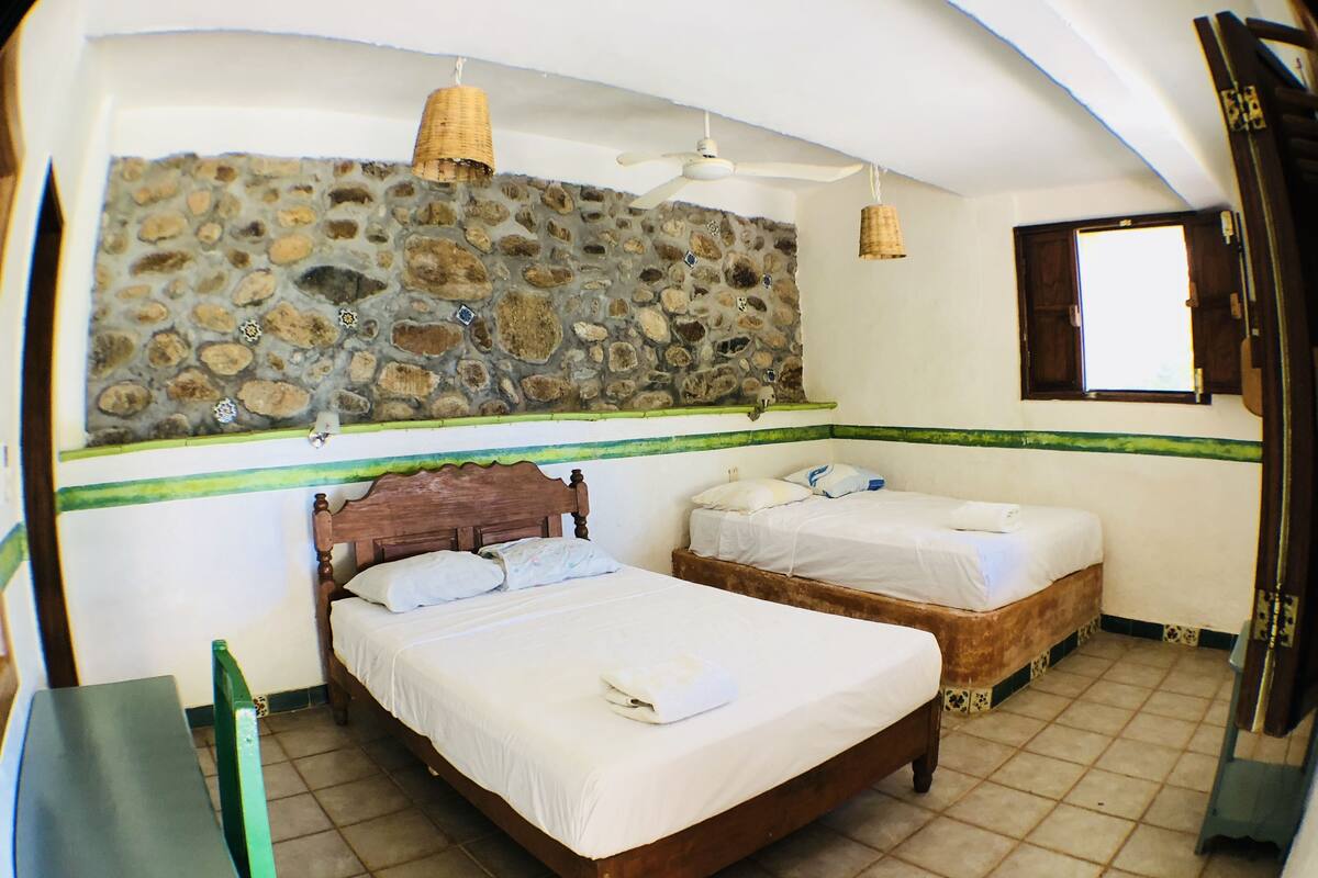 A room with two wooden beds, a stone wall, a ceiling fan, and a window.