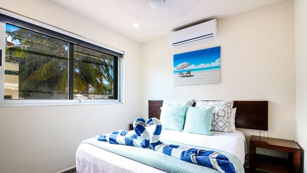 Une chambre à coucher avec un lit, une table de chevet, une fenêtre donnant sur des palmiers, un climatiseur et un tableau représentant une scène de plage.
