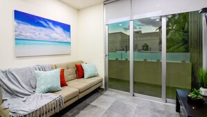 Un salon moderne avec un canapé beige, des coussins à motifs, un grand tableau représentant une plage et une porte-fenêtre coulissante donnant sur un espace extérieur.