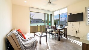 A modern living room with a sofa, a dining table with chairs, a TV mounted on the wall, and a balcony with a view of buildings. - The Furlough Fur204 (Playa del Carmen)