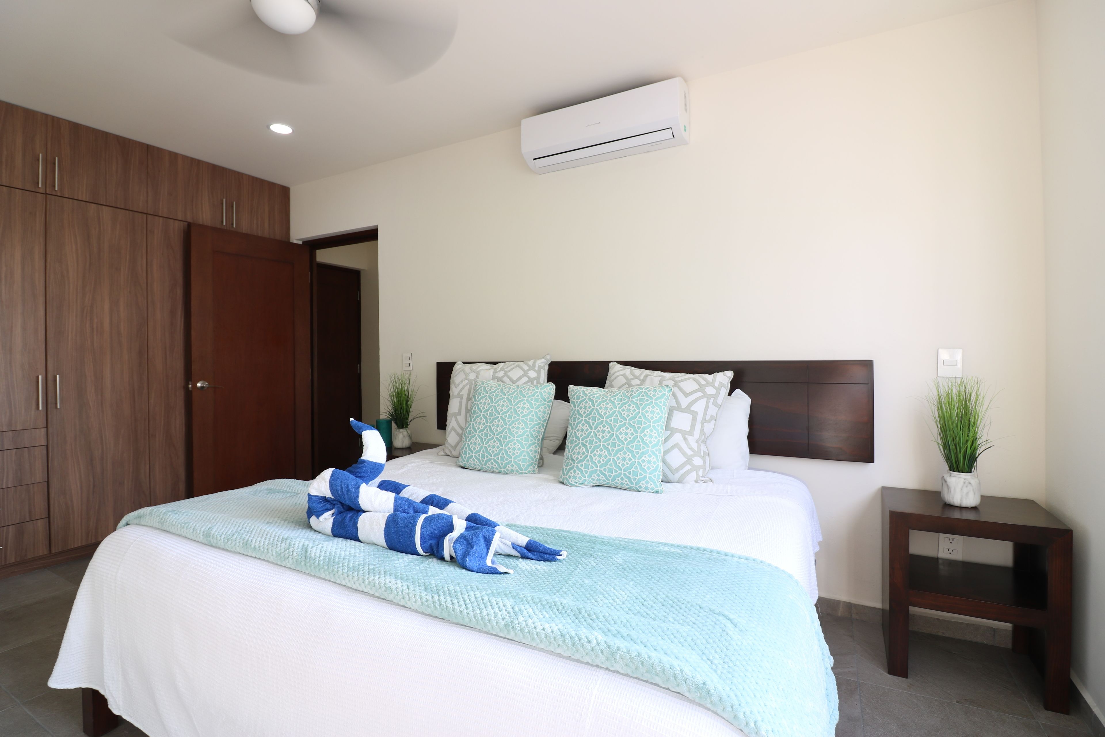 A bedroom with a large bed, a wooden wardrobe, an air conditioner, and a small bedside table with a plant.
