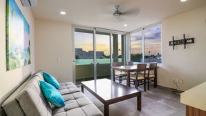 A living room with a sofa, a coffee table, and a dining table with chairs. There is a large window with a view of buildings and a sunset. - The Furlough Fur204 (Playa del Carmen)