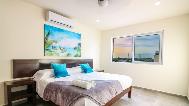 A bedroom with a bed, two nightstands, a large window with a city view, an air conditioning unit, and a painting of a tropical scene. - The Furlough Fur204 (Playa del Carmen)