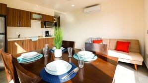 Un coin repas avec une table en bois dressée pour quatre personnes, une cuisine en arrière-plan et un canapé avec des coussins rouges.