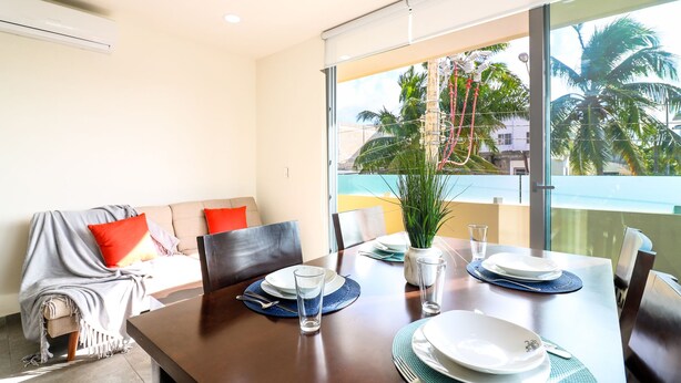 Un coin repas avec une table dressée pour quatre personnes, un canapé avec des coussins et une vue sur des palmiers et un bâtiment, visible à travers une porte vitrée.