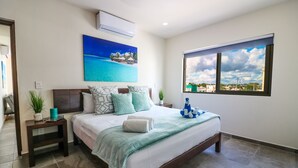 A bedroom with a bed, a nightstand, a potted plant, a painting, and a window with a city view. - The Furlough Fur304 (Playa del Carmen)