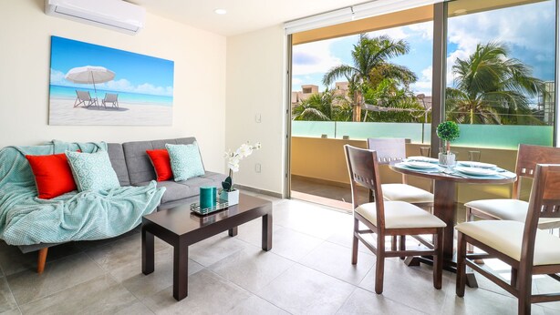 A living room with a sofa, coffee table, and dining table with chairs. There is a painting of a beach scene on the wall, and a balcony with a view of palm trees and buildings. - The Furlough Fur304 (Playa del Carmen)