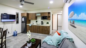 A modern kitchen with wooden cabinets, a built-in microwave, and a refrigerator. There is a dining area with a table and chairs, and a living room with a sofa and a beach-themed wall mural. - The Furlough Fur304 (Playa del Carmen)