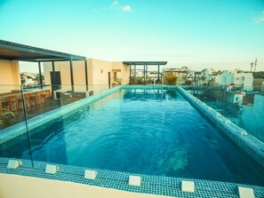 A rooftop infinity pool with clear blue water, glass railings, and a view of the cityscape.