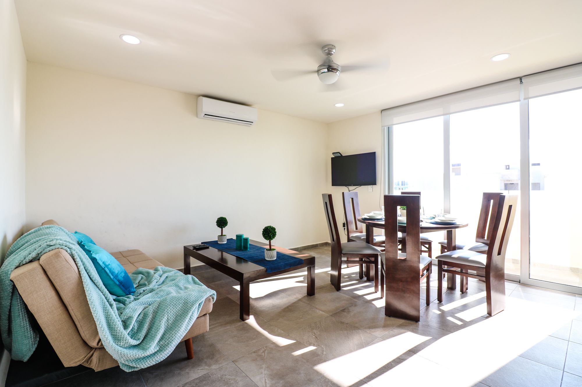 Ein Wohnzimmer mit einer Couch, einem Couchtisch und einem Essbereich mit Stühlen und einem Tisch. Es gibt einen Deckenventilator, einen flachbildfernseher an der Wand und eine Klimaanlage.