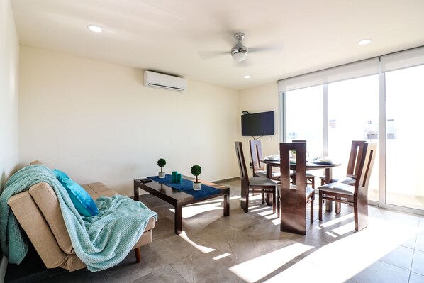 A living room with a sofa, a coffee table, and a dining area with chairs and a table. There is a ceiling fan, a flat-screen TV mounted on the wall, and an air conditioning unit.
