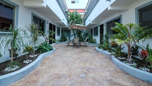 A courtyard with a stone floor, surrounded by planters with various plants, a central table with chairs, and an umbrella. - Apartment Fogon 001 (Playa del Carmen)