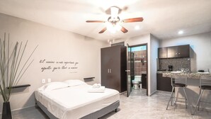 A modern bedroom with a bed, a ceiling fan, a wall quote, a potted plant, and a view into the kitchen. - Apartment Fogon 003 (Playa del Carmen)