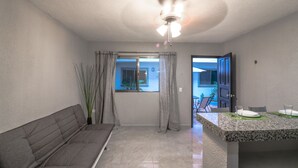 A room with a grey sofa, a ceiling fan, a window with curtains, and a dining area with a table and chairs. - Apartment Fogon 003 (Playa del Carmen)