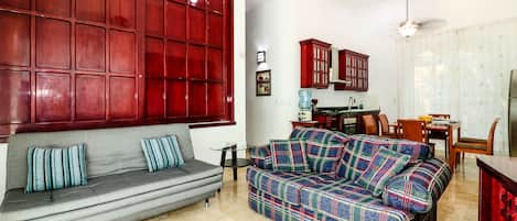 A living room with a grey sofa, a colorful plaid sofa, a small round table, and a kitchen area with wooden cabinets and a refrigerator.